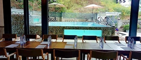 Un área de comedor al aire libre con mesas y sillas, una piscina al fondo y sombrillas.
