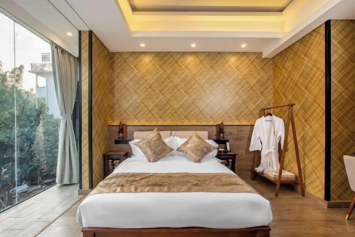 A modern bedroom with a large bed, a wooden headboard, a bedside table, a nightstand, a hanging robe, and a view of greenery outside.