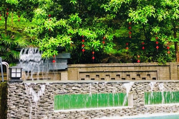 Una cascada fluye sobre una pared de piedra hacia una piscina, rodeada de exuberante follaje y flores rojas.