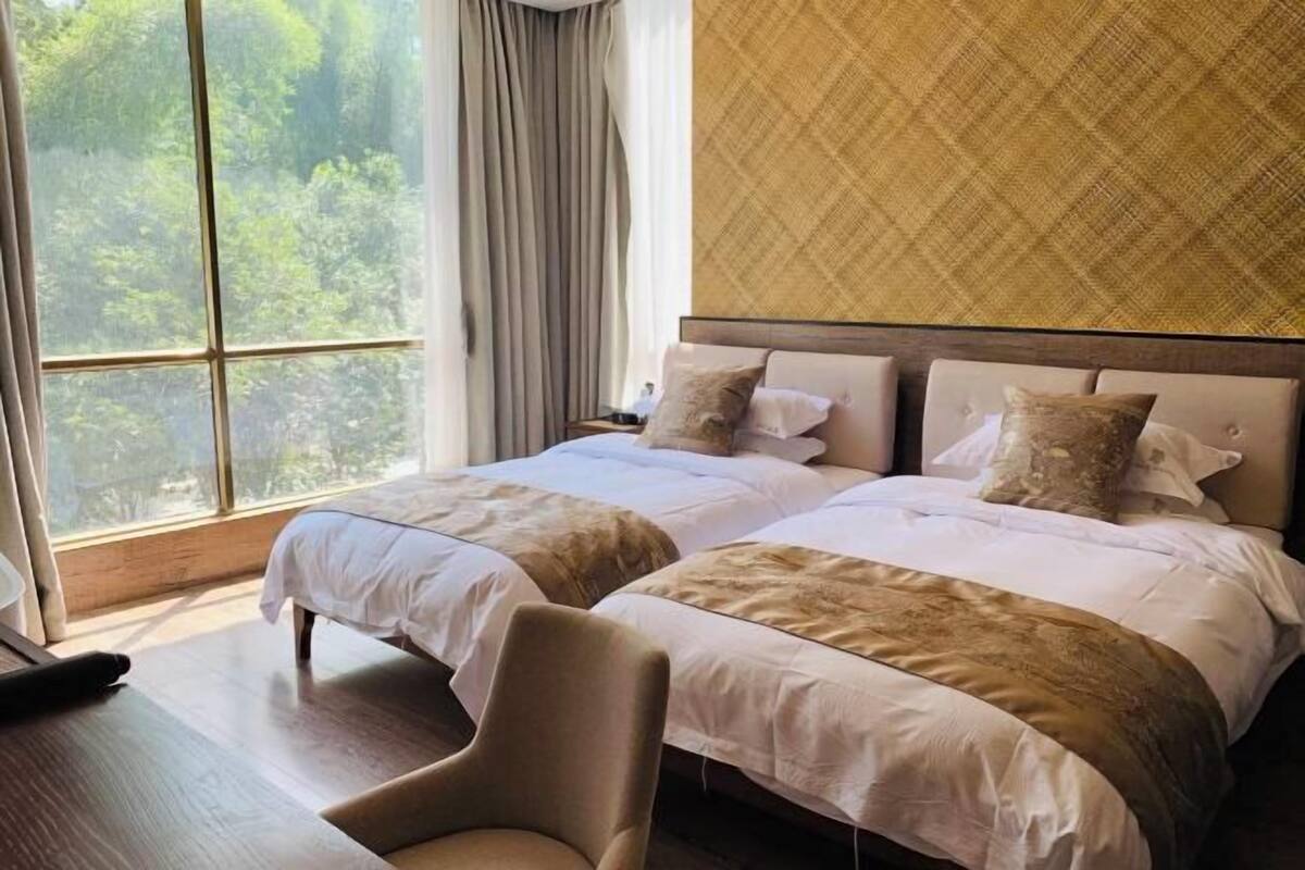 A hotel room with two beds, a wooden floor, a large window with curtains, and a view of greenery outside.