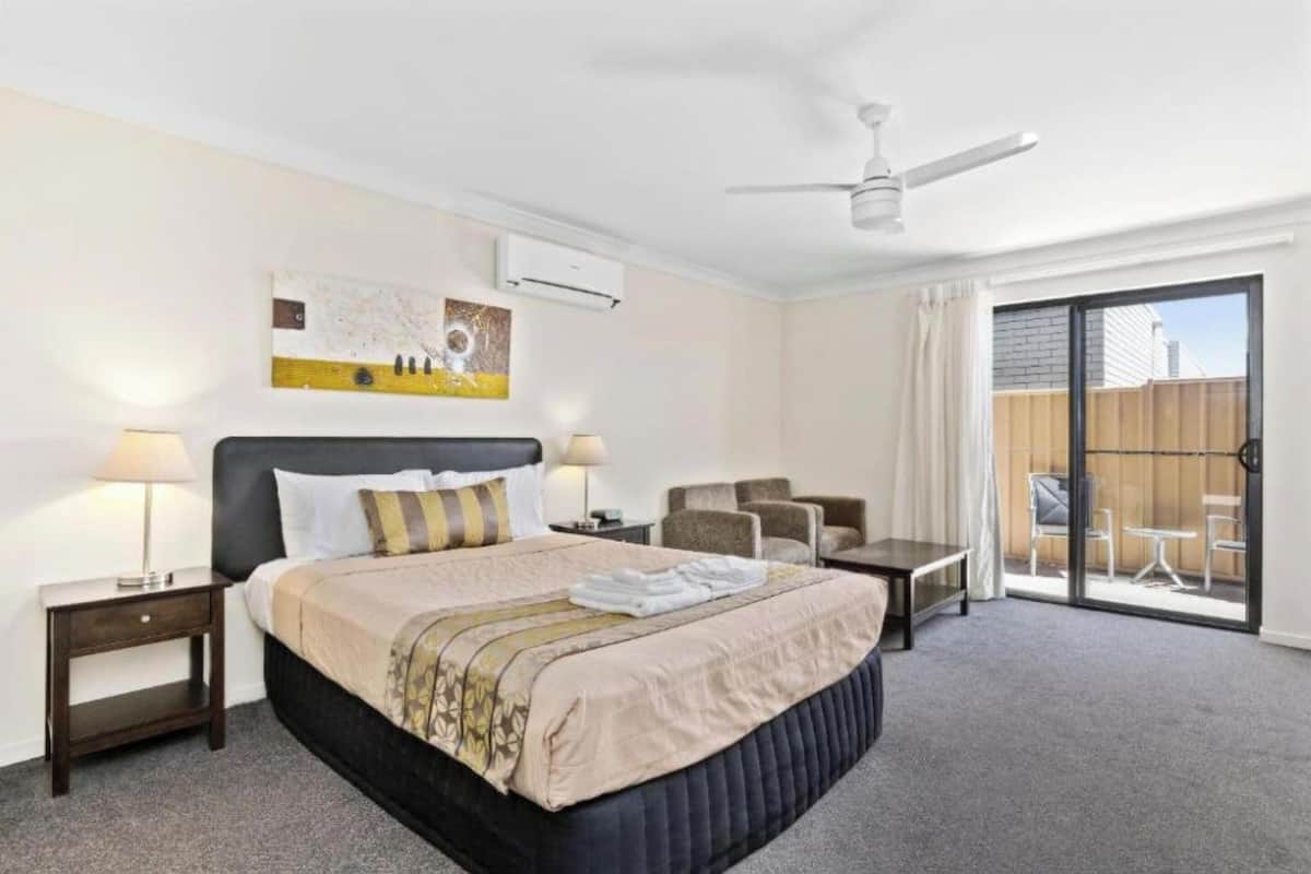 Une chambre d’hôtel avec un grand lit, deux tables de chevet avec des lampes, un ventilateur de plafond, une porte-fenêtre coulissante donnant sur un balcon et un tableau au mur.