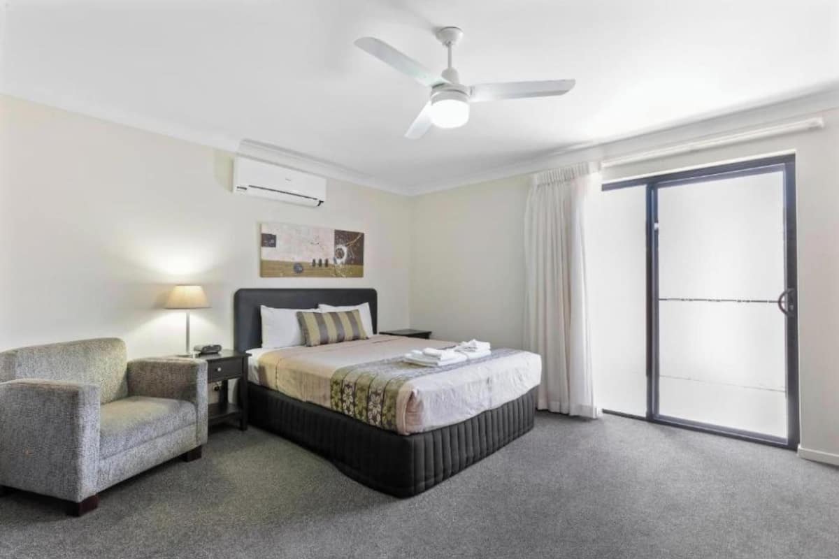 Une chambre d’hôtel comprenant un lit, une chaise, une table de chevet, une lampe, un ventilateur de plafond et une porte-fenêtre coulissante.