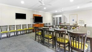 A dining area with a long table and multiple chairs, a television mounted on the wall, and a ceiling fan.