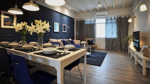 Ein moderner Essbereich mit einem weißen Tisch, blauen Stühlen und einer Vase mit weißen Blumen. Im Hintergrund befinden sich eine Couch und ein Fernseher.