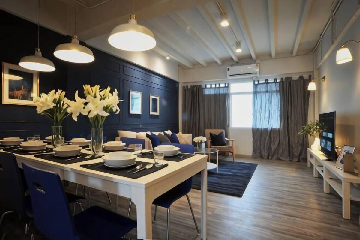 Ein moderner Essbereich mit einem weißen Tisch, blauen Stühlen und einer Vase mit weißen Blumen. Im Hintergrund befinden sich eine Couch und ein Fernseher.