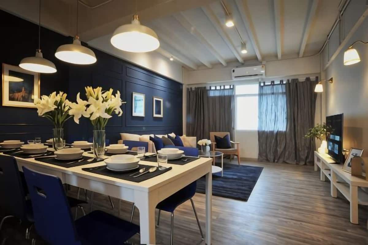 Ein moderner Essbereich mit einem weißen Tisch, blauen Stühlen und einer Vase mit weißen Blumen. Im Hintergrund befinden sich eine Couch und ein Fernseher.