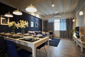 Ein moderner Essbereich mit einem weißen Tisch, blauen Stühlen und einer Vase mit weißen Blumen. Im Hintergrund befinden sich eine Couch und ein Fernseher.