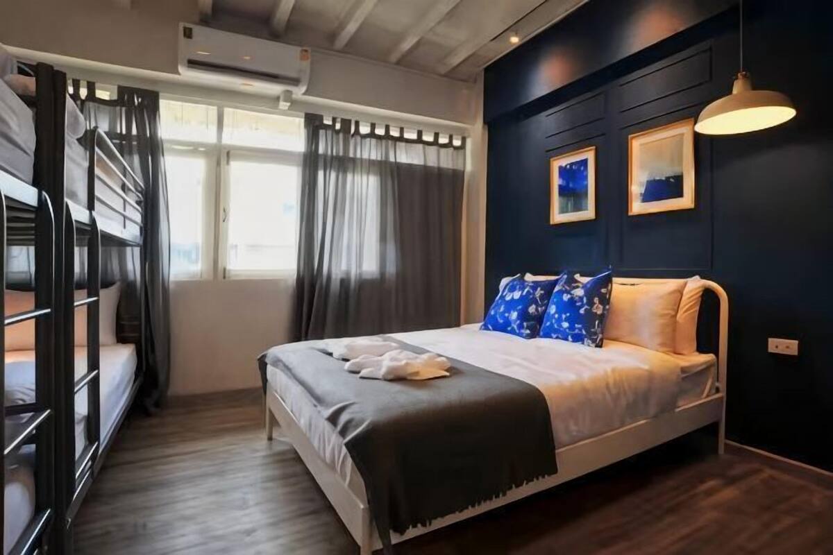 A room with a bunk bed, a single bed, a window with curtains, and a wall-mounted air conditioner.
