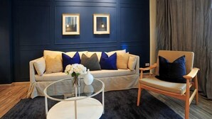 Ein Zimmer mit dunkelblauer Wand, einem Sofa mit gemusterten Kissen, einem runden Couchtisch und einem Sessel.