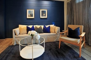 Ein Zimmer mit dunkelblauer Wand, einem Sofa mit gemusterten Kissen, einem runden Couchtisch und einem Sessel.