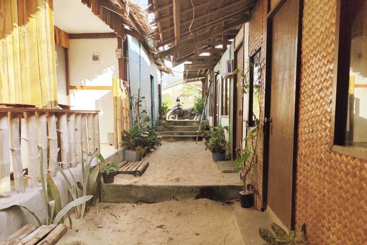 A narrow, sandy pathway flanked by buildings with wooden and woven walls, leading to a staircase.
