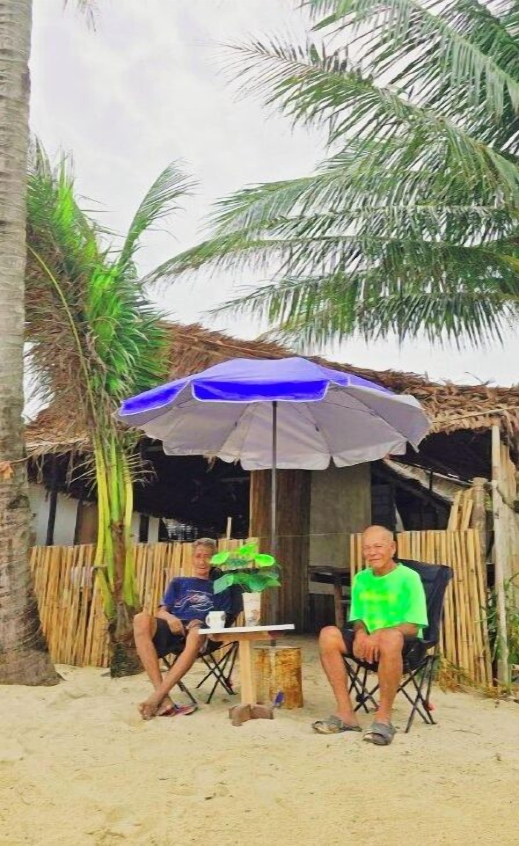 Two people seated under a blue and white umbrella on a sandy surface with palm trees in the background.