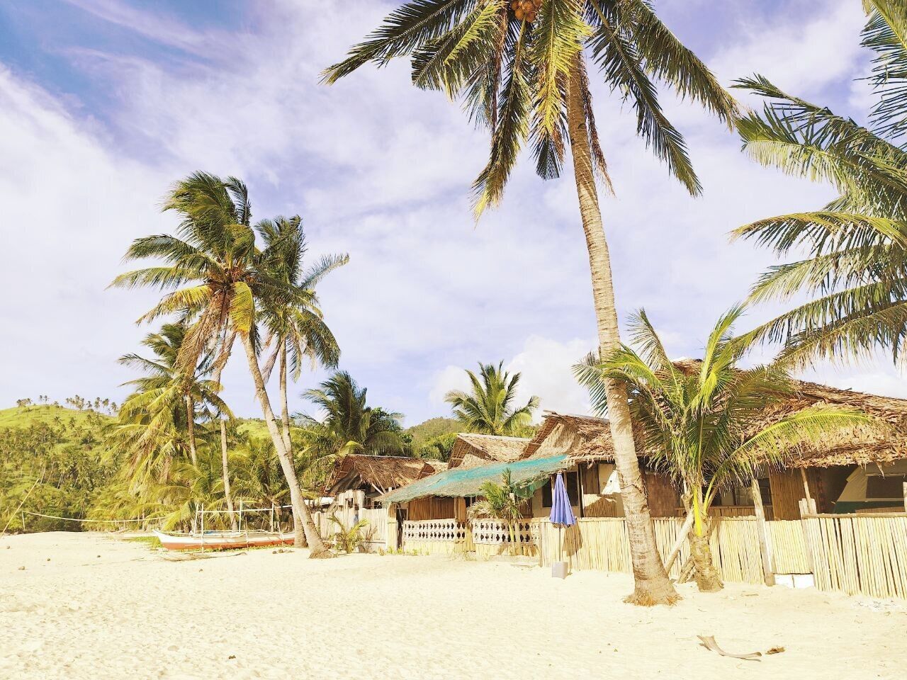 A beach with palm trees, huts, and a boat on the sand.