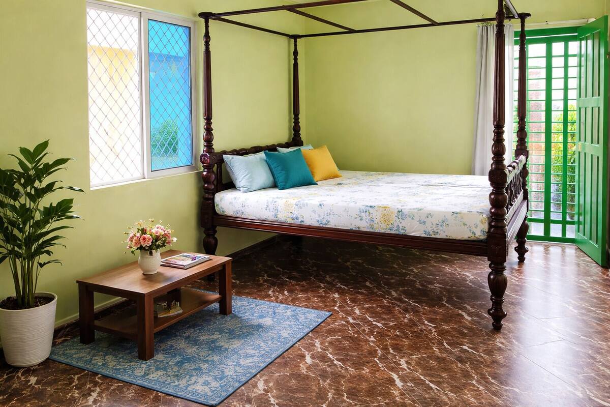 Una cama con dosel adornada con una colcha floral, una mesita de madera con un jarrón de flores, una planta en maceta y un tapete azul estampada.