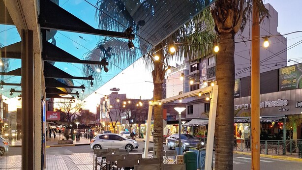 Un café al aire libre con mesas y sillas, suelo de baldosas y vista a la calle con coches y edificios.
