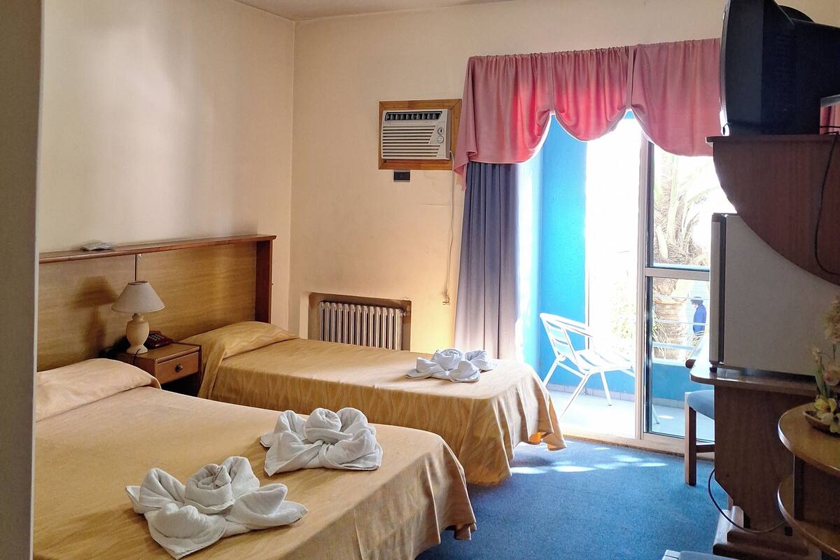 Habitación de hotel con dos camas, un escritorio con televisor, una ventana con cortinas y un radiador.