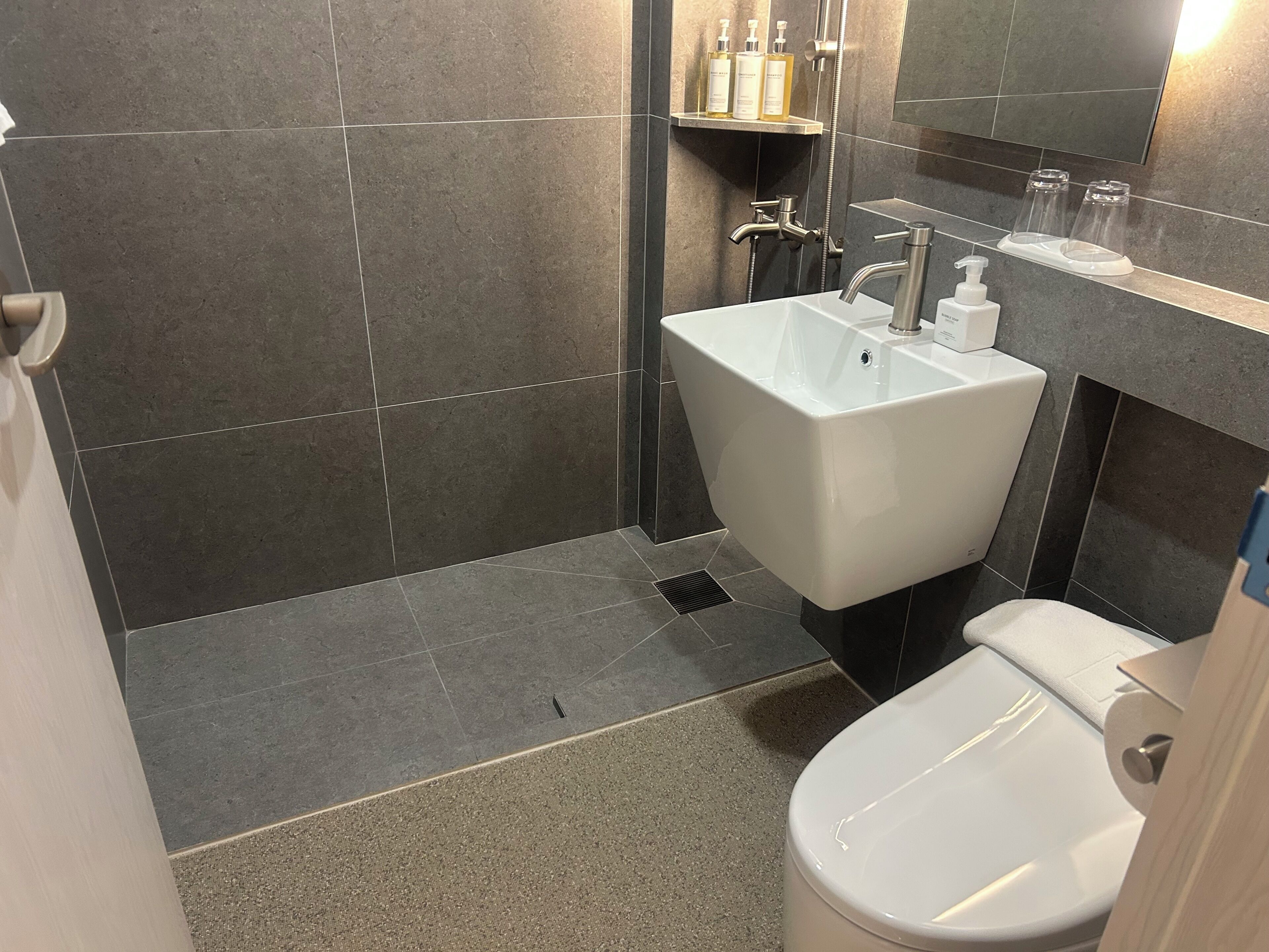 Une salle de bain avec un lavabo blanc, une douche et des toilettes. On trouve des articles de toilette sur une étagère et un miroir au-dessus du lavabo.