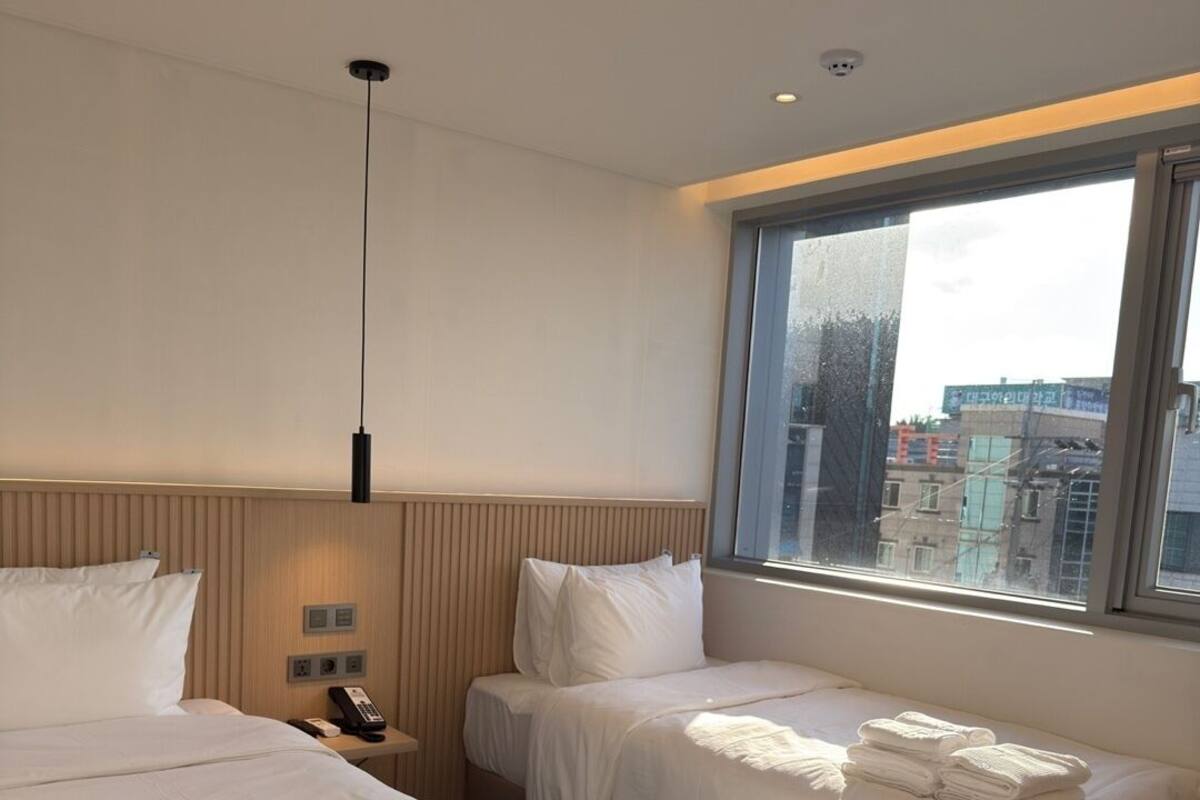 A hotel room with two beds, a bedside table with a phone, a window with a city view, and a hanging light fixture.