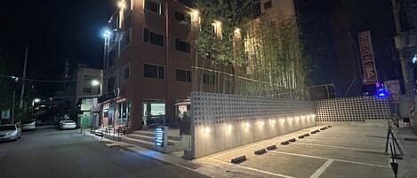 A building with multiple floors, a parking area with marked spaces, and a fence with illuminated panels.