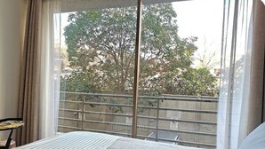 Une chambre d’hôtel avec un lit, une fenêtre donnant sur des arbres et des immeubles, et un balcon avec une balustrade.