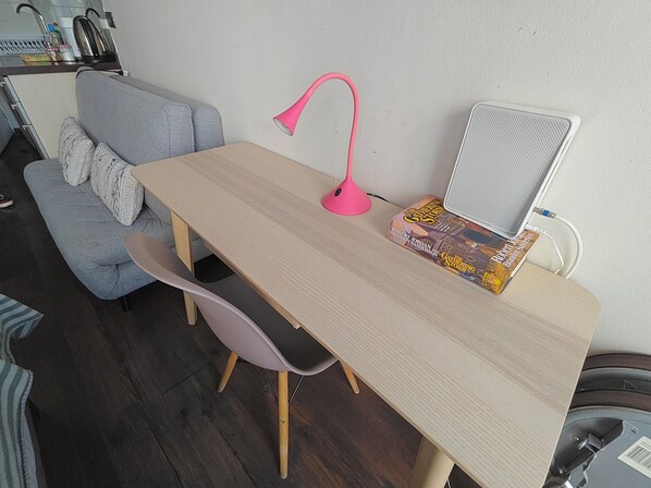 Une petite pièce chaleureuse, avec une table en bois, une lampe de bureau rose, une chaise, un canapé gris et une bibliothèque remplie de livres.