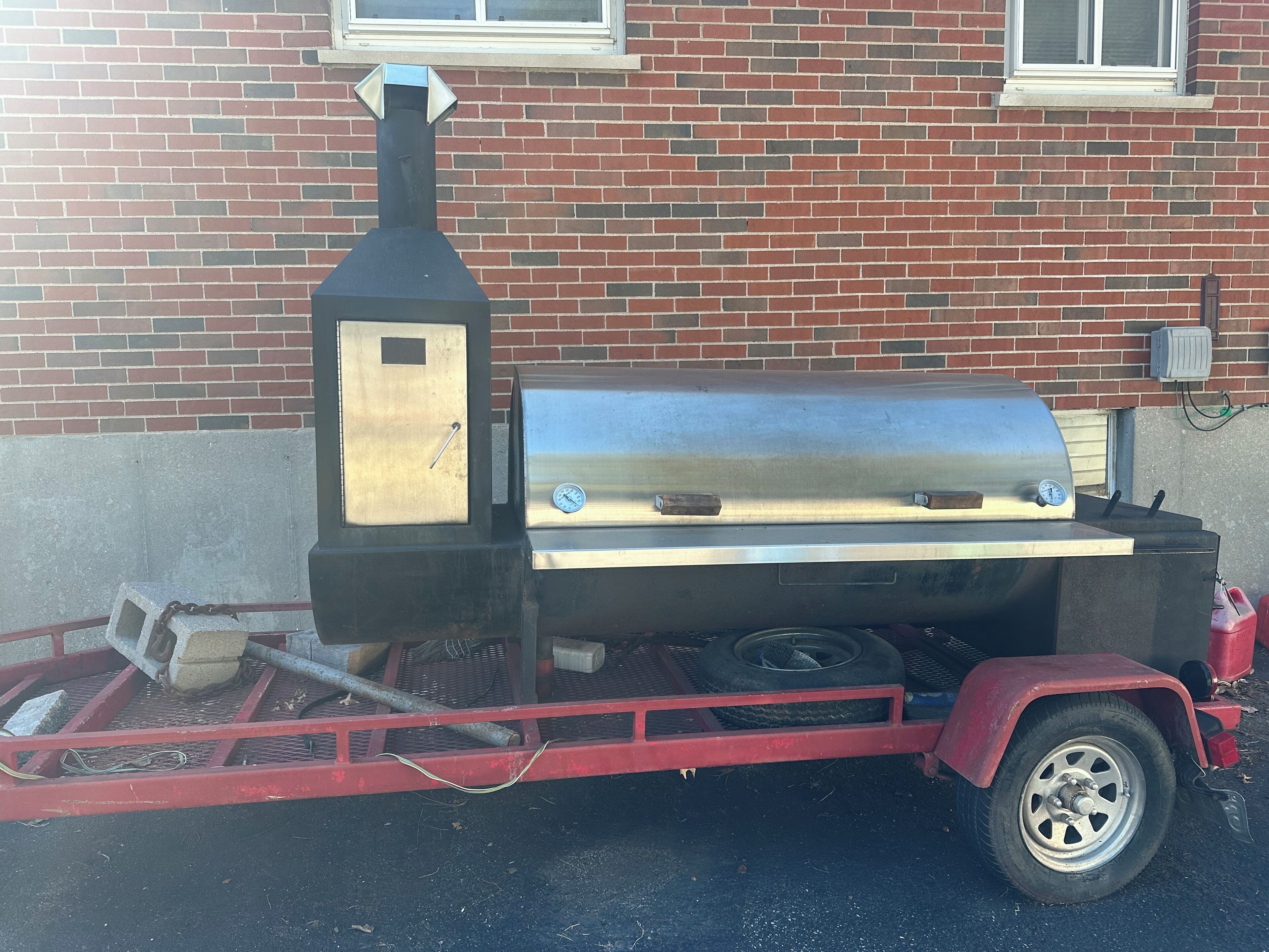 A portable barbecue grill with a chimney, a metal lid, and a side burner, mounted on a red trailer.