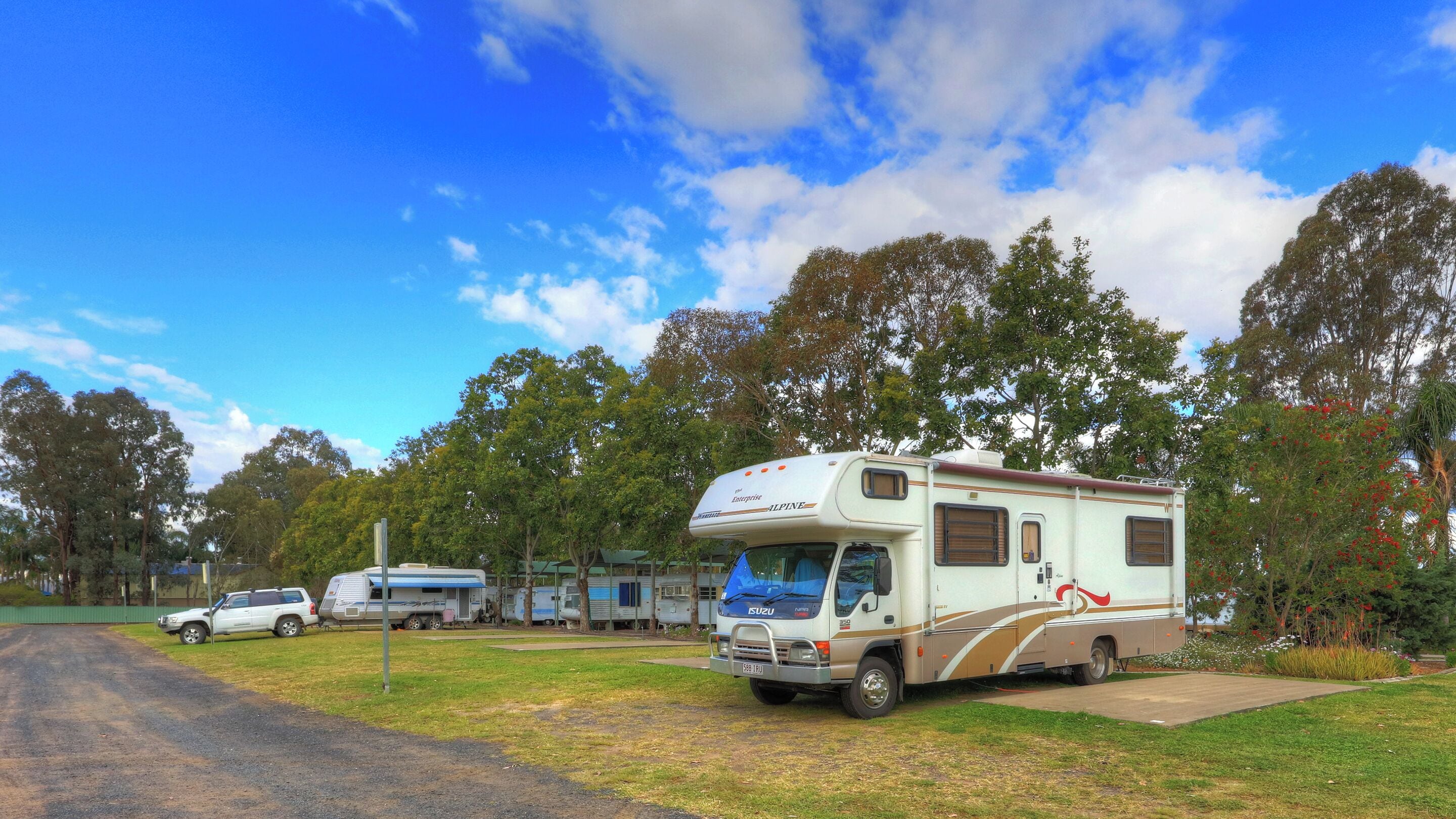 A caravan park with a large Isuzu camper parked on a grassy area, surrounded by trees and other caravans.