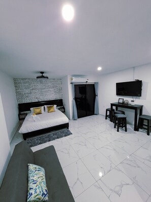 A modern hotel room with a bed, a sofa, a desk with chairs, a TV, and a ceiling fan.