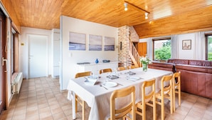 Uma sala de jantar com uma mesa de madeira posta para seis pessoas, cercada por cadeiras de madeira. Uma lareira de tijolos é visível ao fundo, e há uma escada que leva a um andar superior.