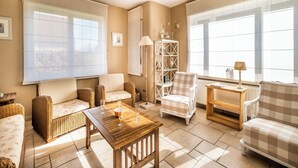 A cozy living room with wicker furniture, a wooden coffee table, and large windows with blinds.