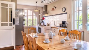 Une cuisine avec une table à manger en bois, préparée pour le thé, un réfrigérateur et une fenêtre donnant sur un paysage verdoyant.