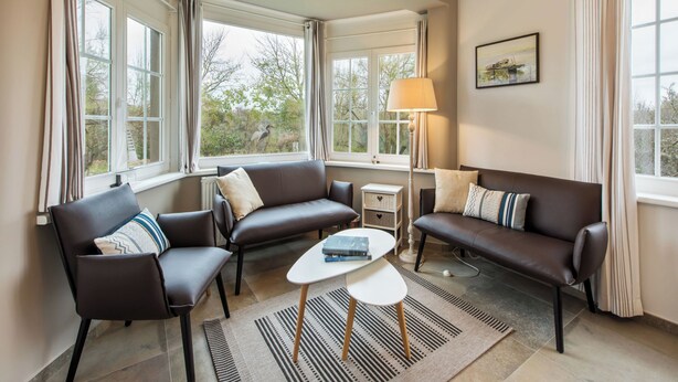 A modern living room with leather sofas, a coffee table, and a lamp. Large windows offer views of trees and a building outside. - Charming villa located by the dunes (Koksijde)