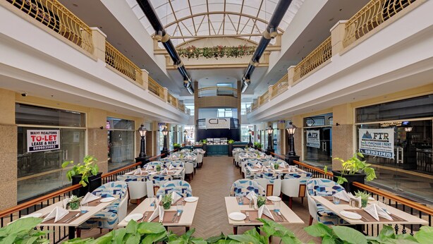 A spacious indoor dining area with multiple tables and chairs, a central open space, and a high ceiling with decorative elements. - AR Suites Sohna Road (Gurugram)