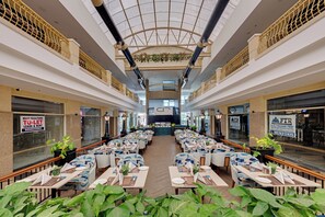 A spacious indoor dining area with multiple tables and chairs, a central open space, and a high ceiling with decorative elements. - AR Suites Sohna Road (Gurugram)