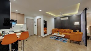 A modern living room with a sofa, coffee table, and orange chairs. There is a kitchen area with a stove and cabinets. - AR Suites Sohna Road (Gurugram)