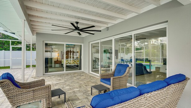 Ein überdachter Patio-Bereich mit Korbmöbeln, einem Deckenventilator und einer Glastür, die zu einem Poolbereich führt.