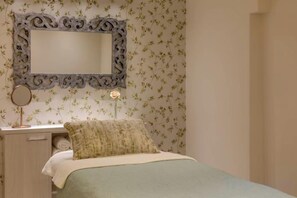 Une chambre à coucher comprenant un lit, une commode, un miroir et un mur recouvert d’un papier peint à motifs floraux.