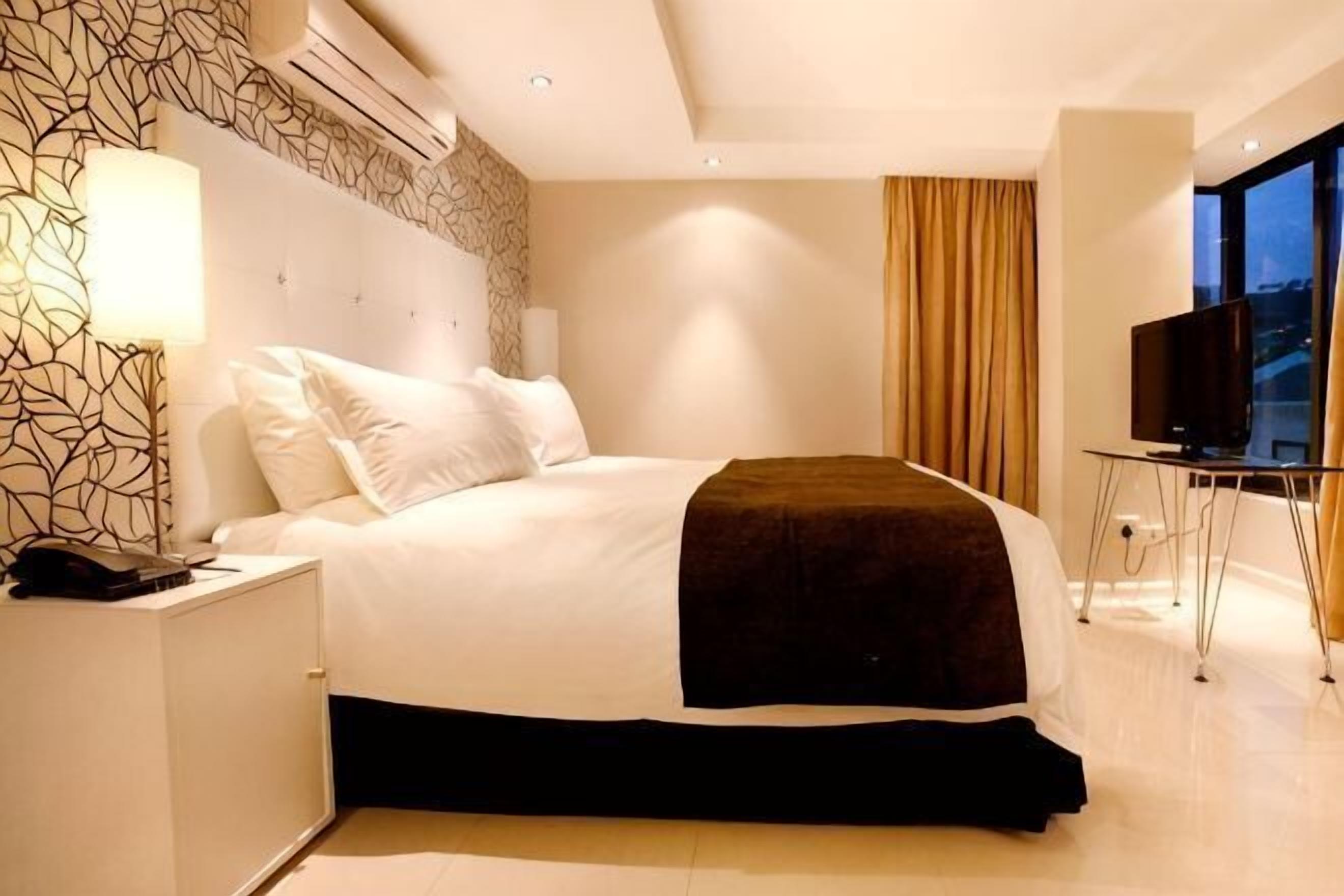 Habitación de hotel moderna con una cama grande, una mesita de noche con lámpara, televisor y vistas a la ciudad.