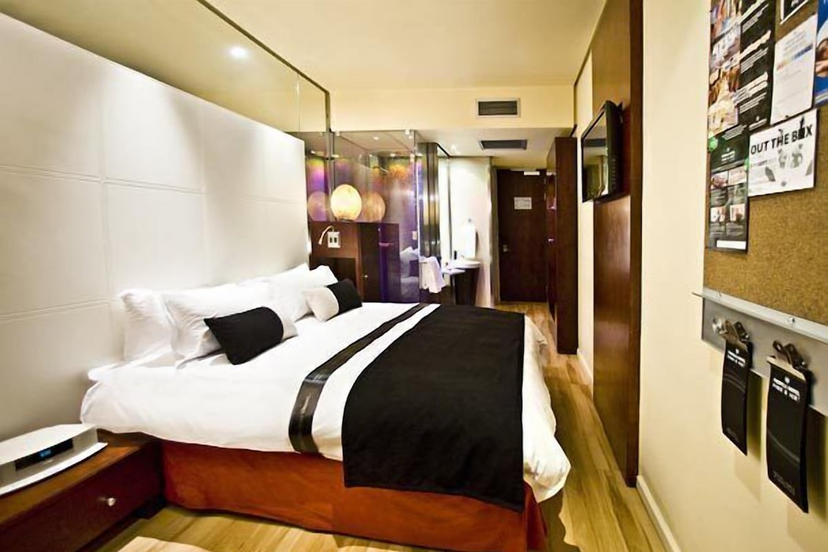 Ein Hotelzimmer mit einem großen Bett, einem Nachttisch, einem Wand-Fernseher und einem Korkbrett mit verschiedenen Aushängen.