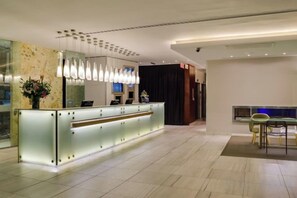 Zona de recepción de hotel con un mostrador moderno, iluminación colgante y un área de estar con sillas.