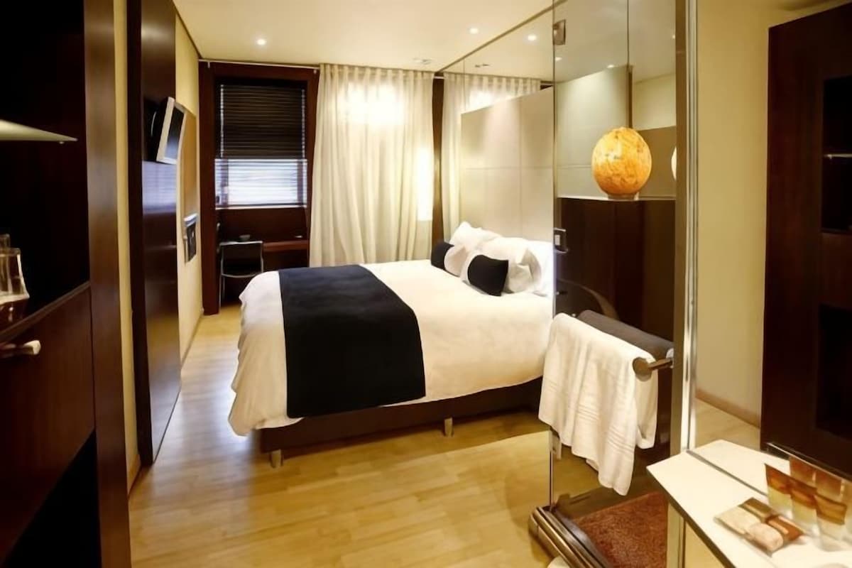 A hotel room with a large bed, a bedside table, a mirror, a TV, and a window with curtains.