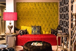 Una habitación con cabecera capitonada, un sofá rojo, un papel tapiz estampado y una pequeña mesa redonda con una lámpara.