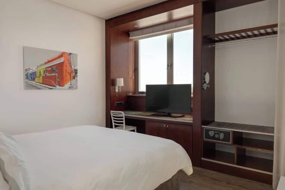A hotel room with a bed, a desk with a television, a wardrobe, and a painting on the wall.