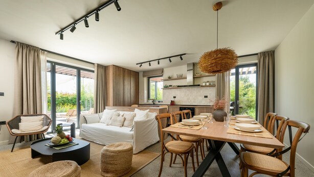 A modern living room with a dining area, large windows, and a kitchen in the background.