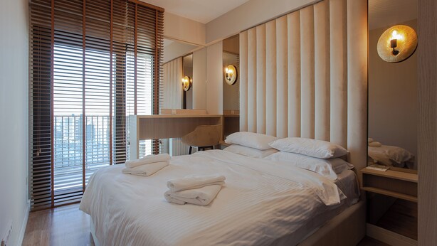 A modern hotel room with a large bed, a bedside table, a mirror, and a view through the window with blinds. - Apartment Skopje Diamond  View (Skopje)