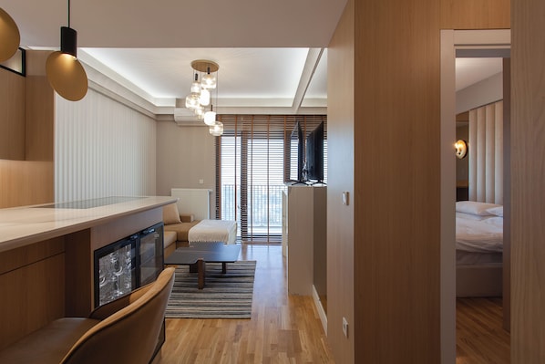 A modern hotel room with a wooden floor, a sofa, a dining table, and a TV in the background.