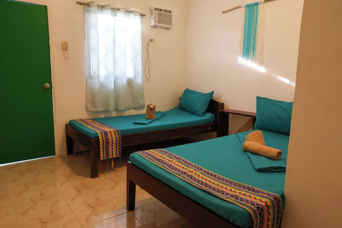 A room with two single beds, each with a teal mattress and a colorful blanket. There is a wooden bench with a cushion and a small table with a towel. A green door and a window with a curtain are visible.