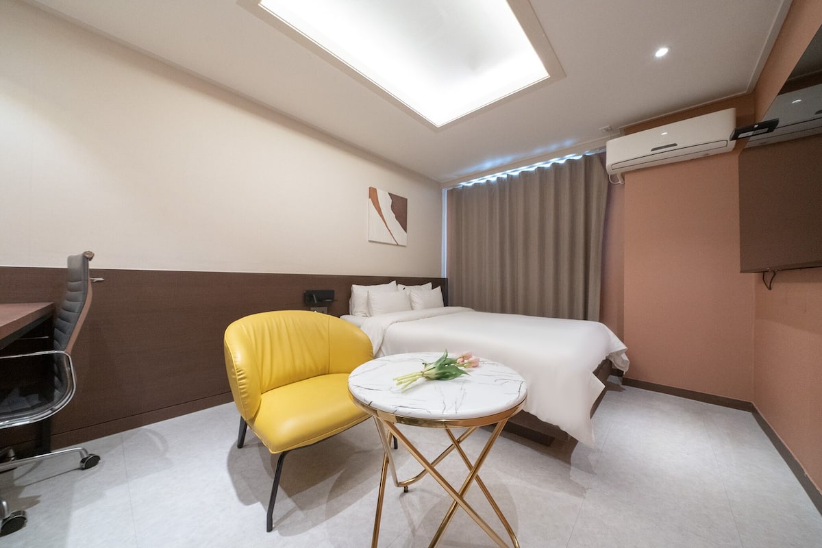 Une chambre d’hôtel avec un lit, une chaise jaune, une petite table ronde ornée de fleurs, un bureau et un appareil de climatisation.