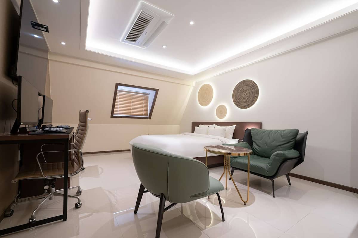 Une chambre d’hôtel moderne, équipée d’un lit, d’un bureau avec un ordinateur, d’un fauteuil vert et d’une petite table ronde.
