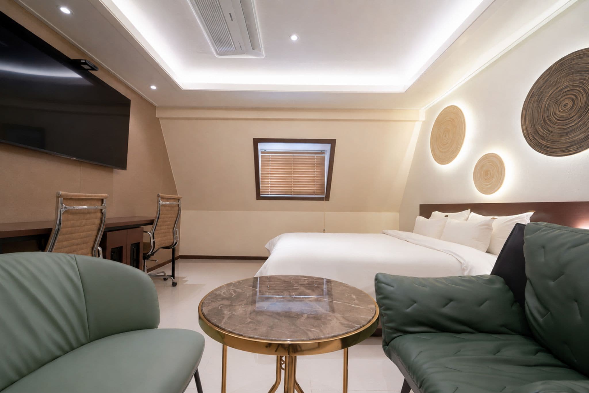 A hotel room with a bed, a round table, two green sofas, a desk with a chair, and a TV mounted on the wall.